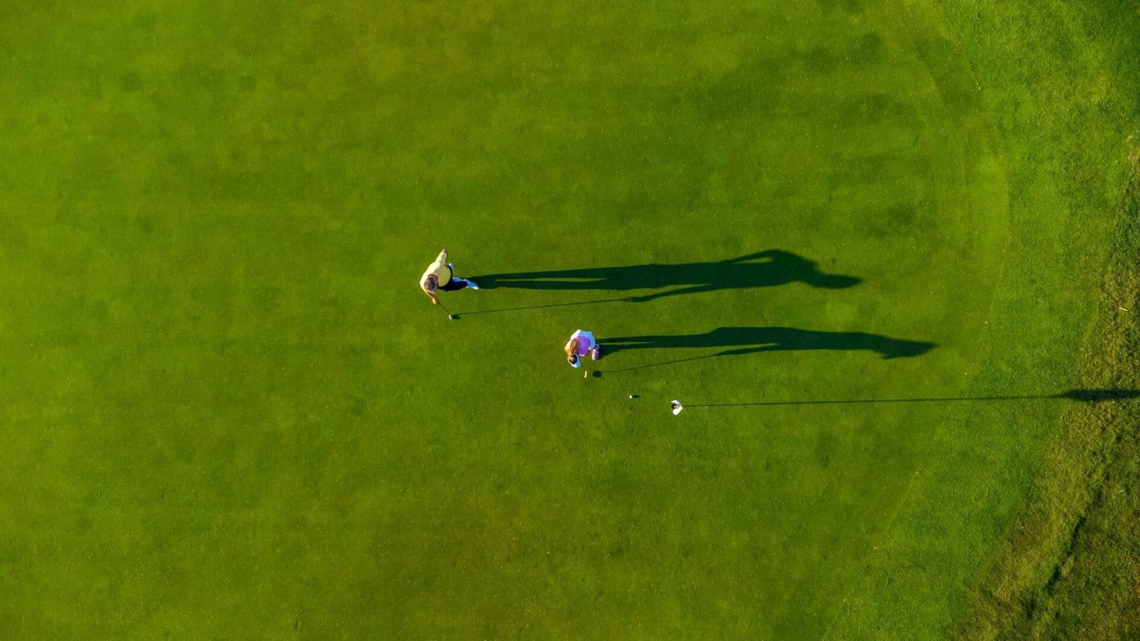 Luftaufnahme von zwei Personen beim Putten auf einem Golfgrün mit langen Schatten im Abendlicht. 